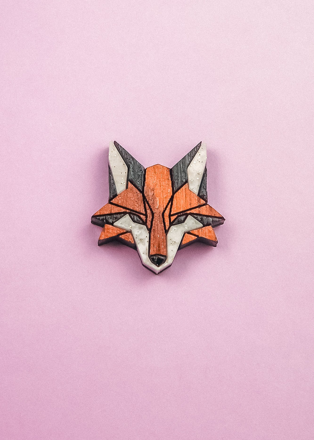 A geometric Fox Pin brooch, crafted from recycled wood in orange, black, and white hues, is displayed on a pink background—perfect for an eco-conscious style.