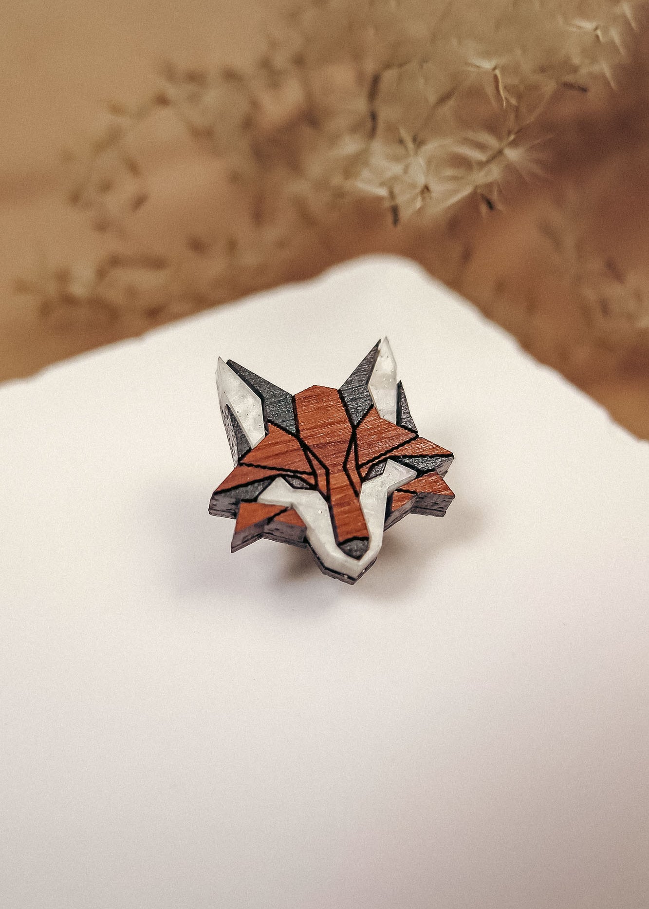A geometric, origami-style handcrafted fox pin made of recycled wood rests on a white surface, with soft beige plant stems blurred in the background—an eco-conscious accessory with natural charm.