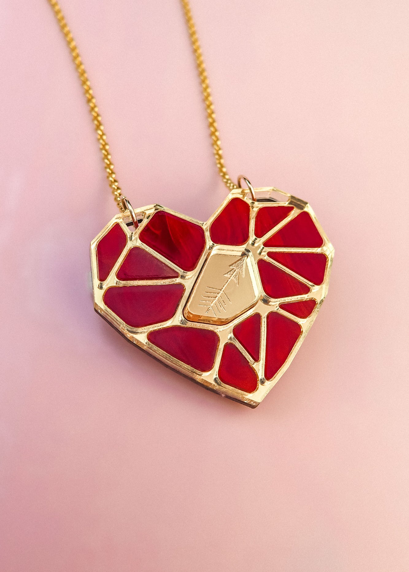 A handcrafted gold chain necklace featuring the Fragment of Love Necklace’s polygon heart pendant, formed from red stained glass outlined in gold, with a central gold plate etched with an arrow, set against a soft pink background.