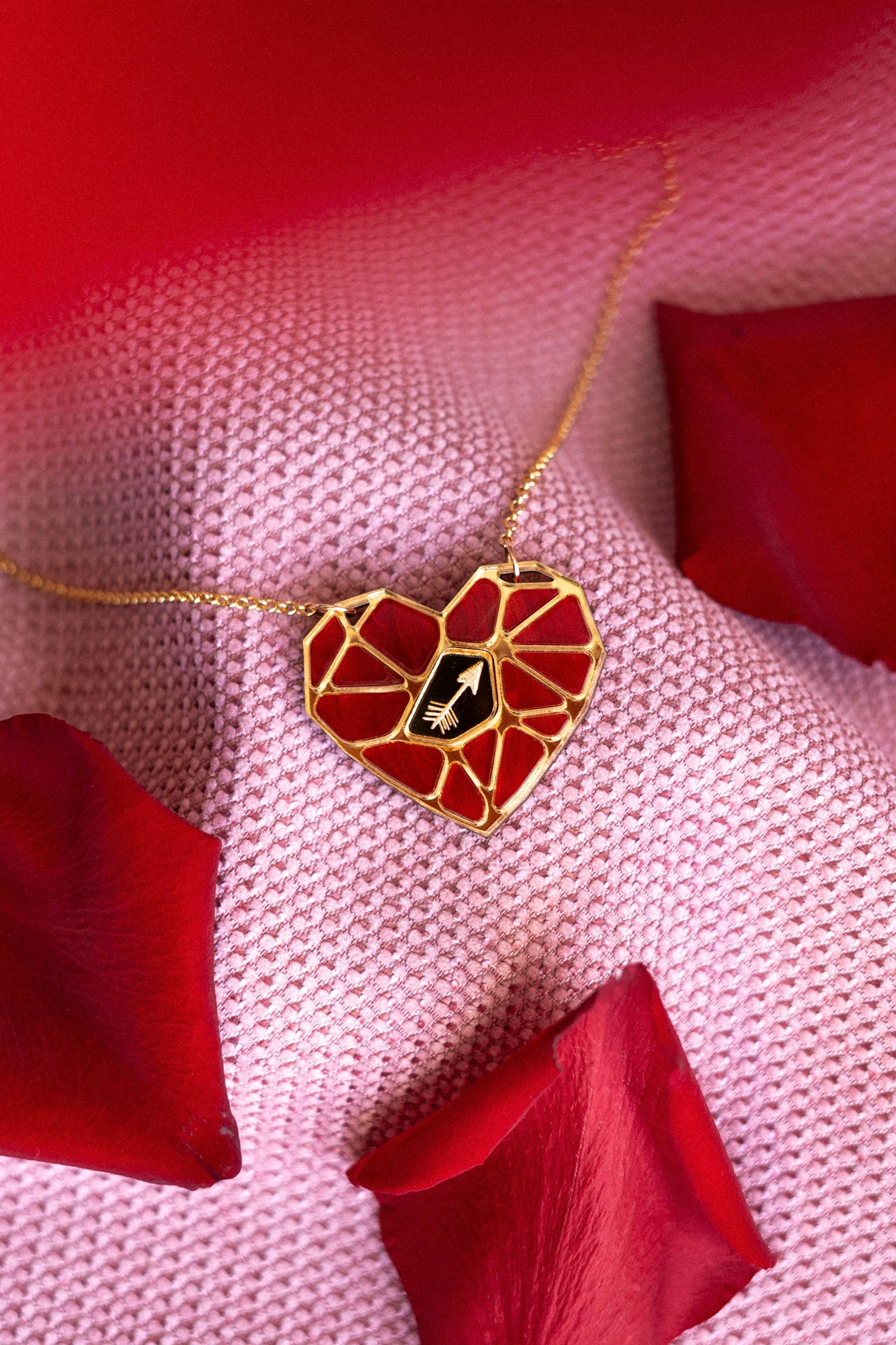 A handcrafted gold heart pendant, featuring a red mosaic design and a small white hand symbol, rests on pink textured fabric amid red rose petals—a striking Fragment of Love Necklace.