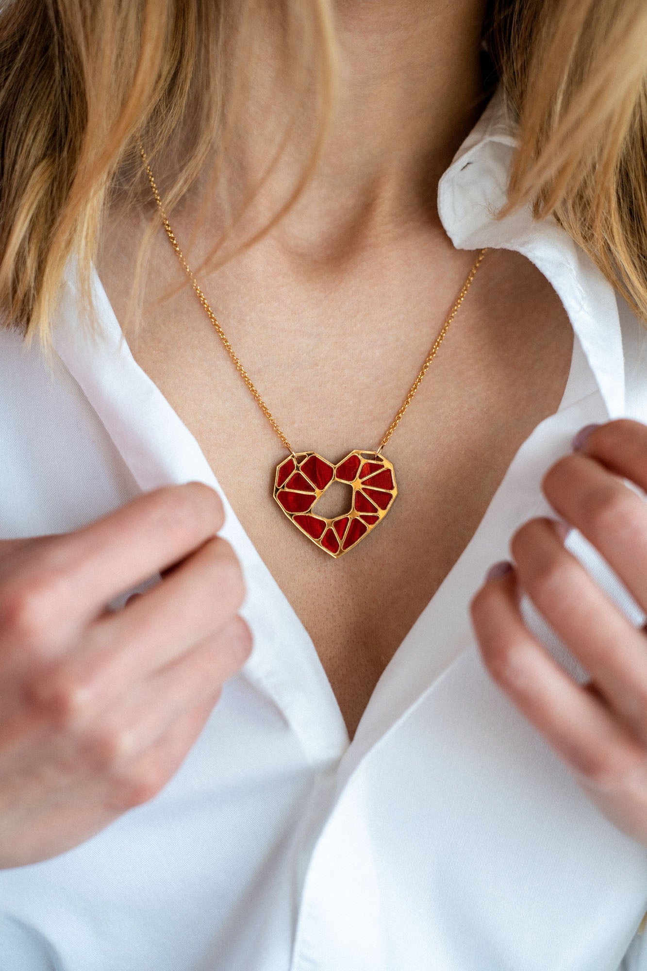 A close-up of a person in a white shirt, holding the collar open to reveal a handcrafted gold necklace with a geometric red heart pendant—an elegant Fragment of Love Necklace displayed around their neck.