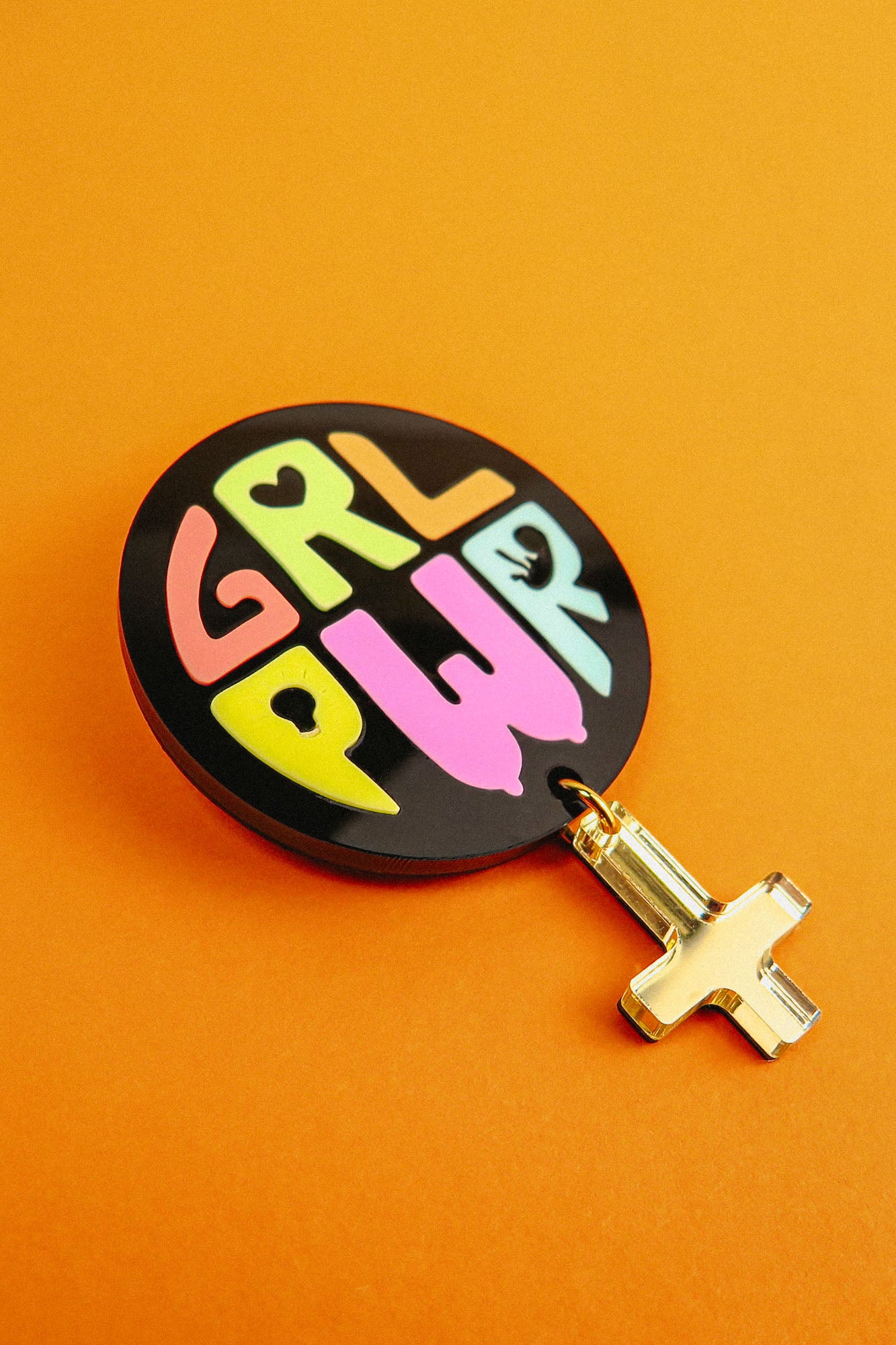 A round black keychain with GRL PWR written in colorful, playful letters, attached to a gold gender symbol charm, lies on a bright orange surface—a handcrafted accessory full of meaningful symbolism.