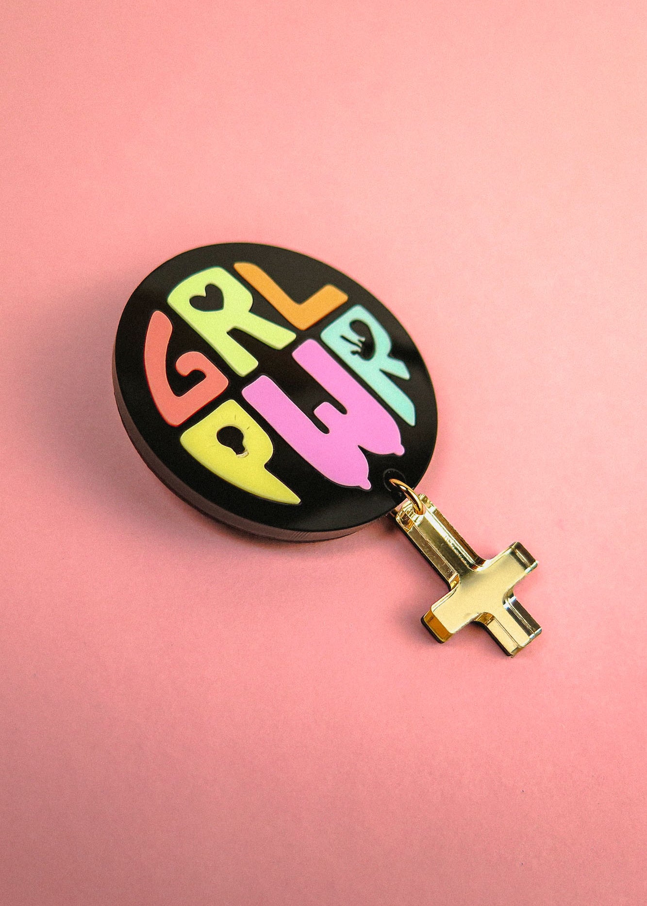 A round black Girl Power Brooch with GRL PWR in colorful letters and a gold female gender symbol charm hanging below, placed on a pink background—a handcrafted accessory celebrating meaningful symbolism.