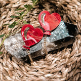 Two red Heart Balloon Studs with gold accents and playful dangling chains are displayed on a large crystal, resting on a woven, natural fiber surface with a small green plant nearby.