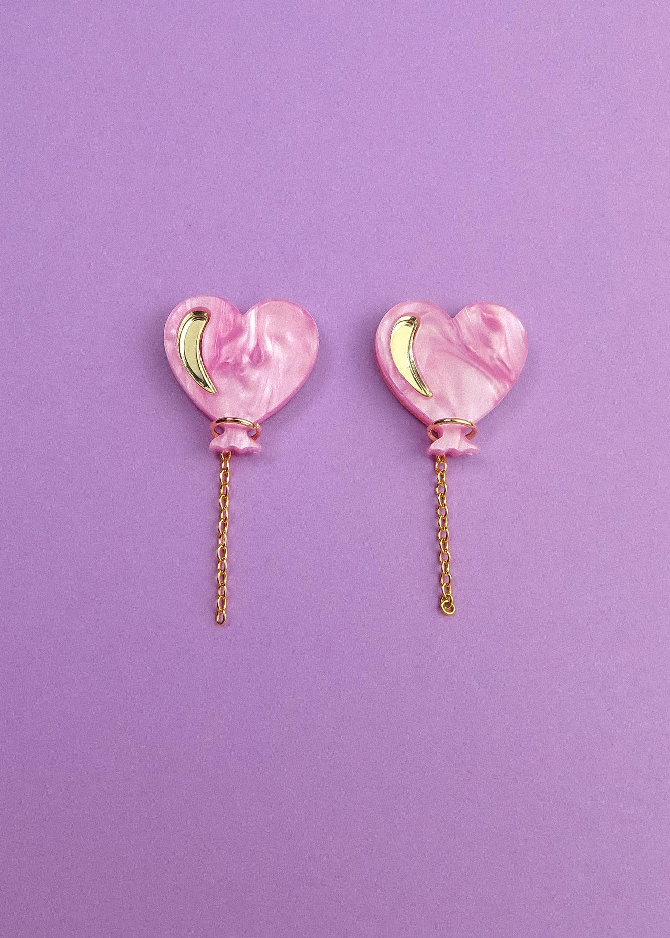 A pair of Heart Balloon Studs—pink, heart-shaped earrings with gold crescent accents and dangling gold chains—adds vibrant style, displayed on a purple background.