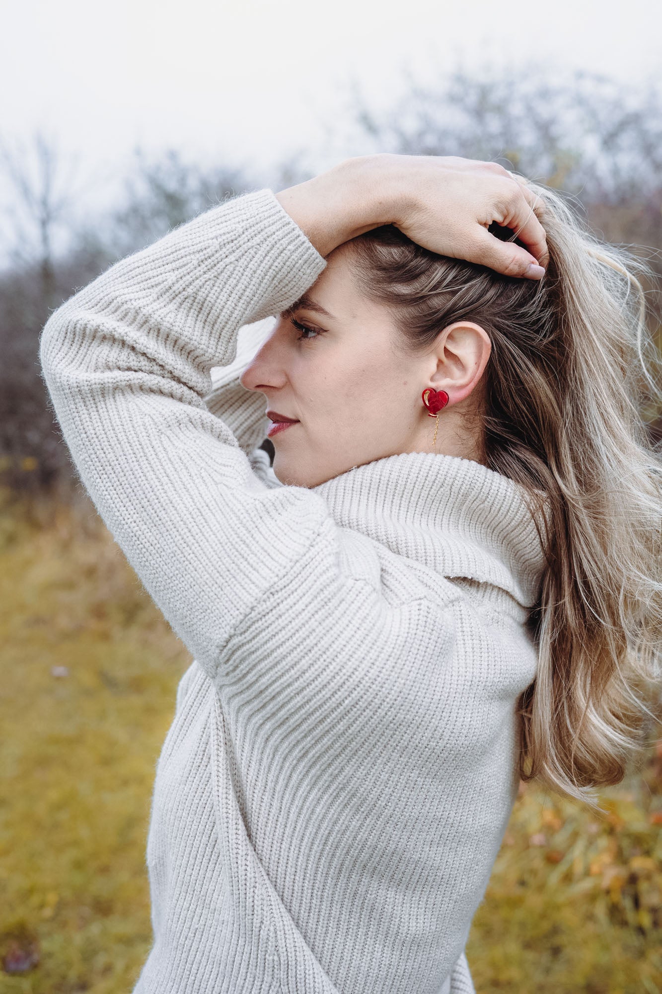 A woman in a cream-colored sweater stands outdoors, facing sideways with one hand holding back her long blonde hair. She wears playful Heart Balloon Studs, adding a romantic jewelry touch to her look. The background is blurry with trees and grass.