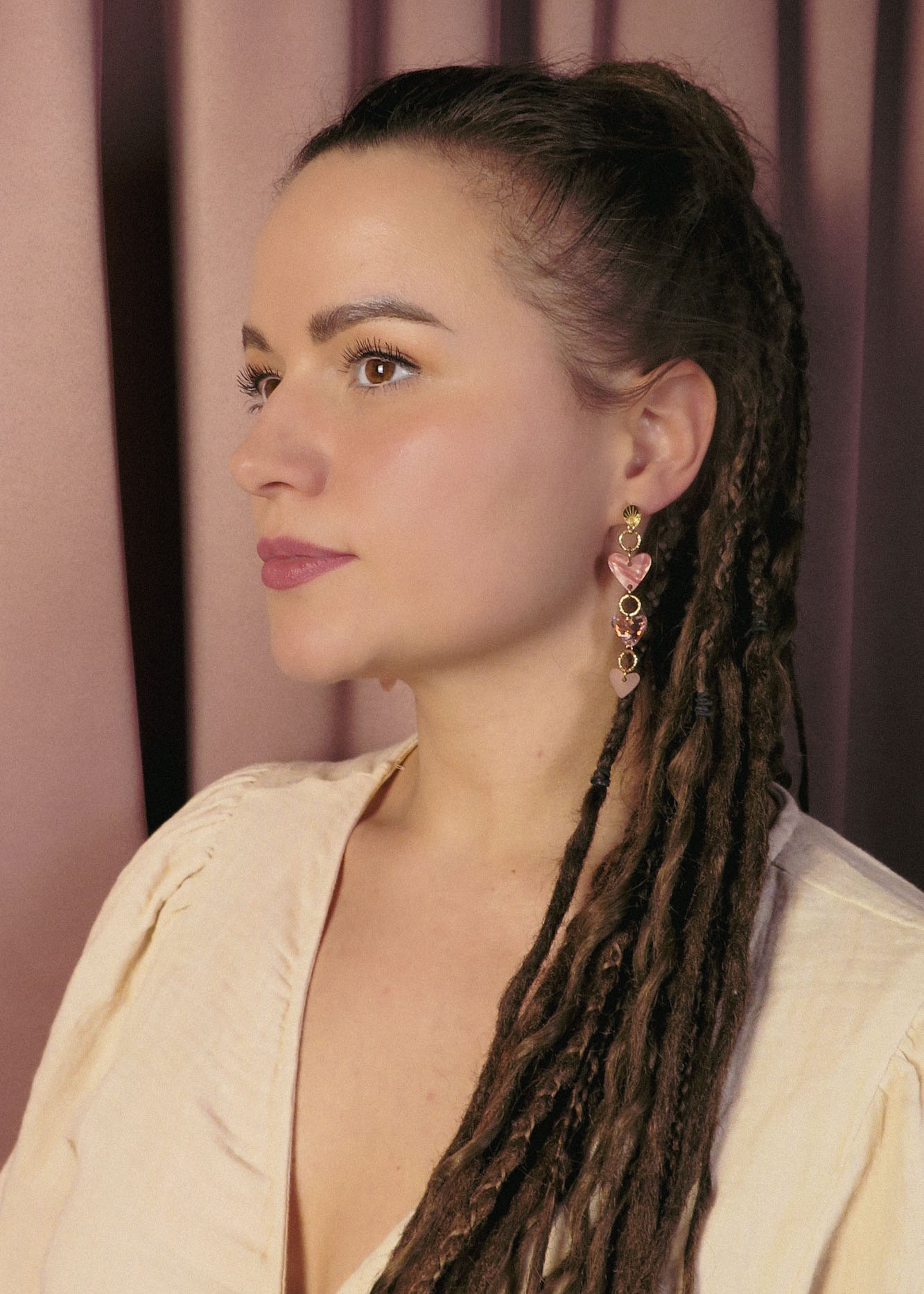 A woman with long brown dreadlocks and gold Heart Chain Earrings gazes to the left. She wears a cream-colored blouse and stands against a backdrop of pink curtains, highlighting soft pink tones throughout the scene.