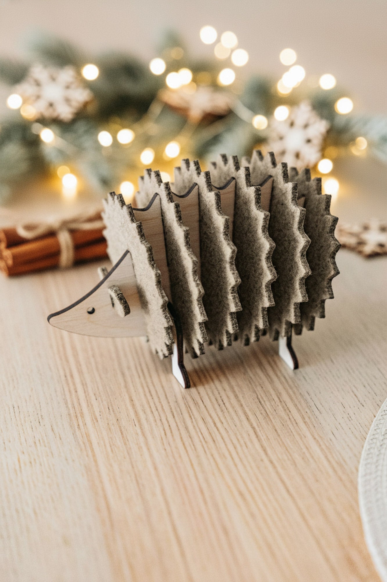A wooden hedgehog figurine, perfect for home decor, sits on a light wooden table. In the blurred background, lights, fir branches, and cinnamon sticks create a festive, cozy atmosphere.
