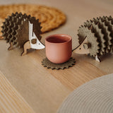 Two wooden hedgehog-shaped felt coasters hold a round, pink cup on a wooden table with woven placemats in the background—a charming touch of home decor.