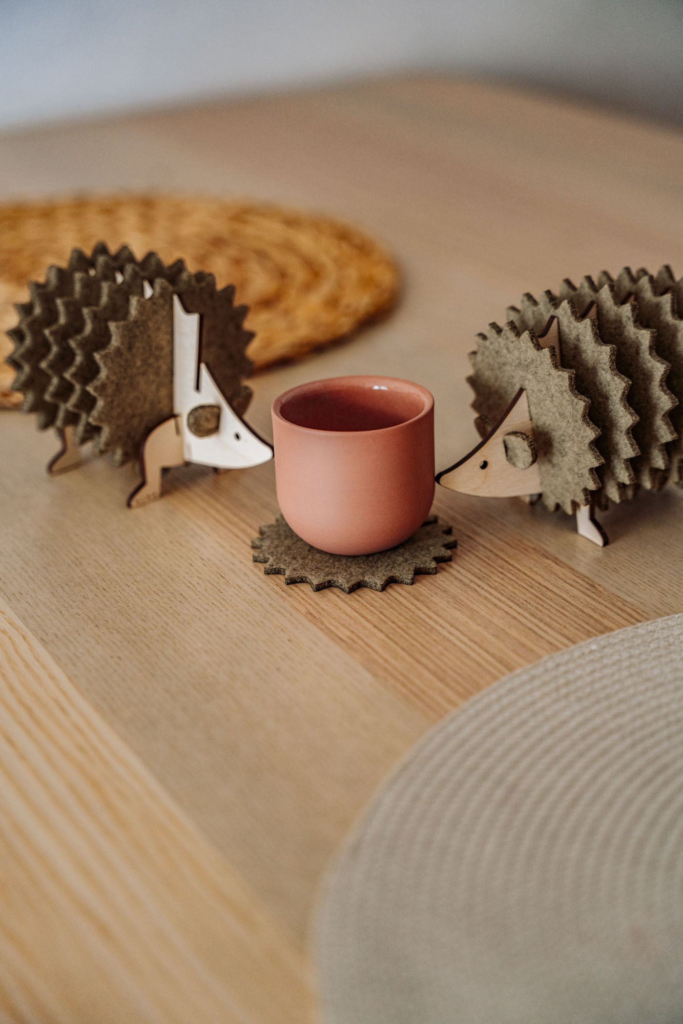 Two wooden hedgehog-shaped felt coasters hold a round, pink cup on a wooden table with woven placemats in the background—a charming touch of home decor.