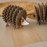 A small, decorative wooden hedgehog coaster holder made from layered, textured materials stands on a light wooden surface, with another similar figurine blurred in the background—perfect for adding charm to your home decor.