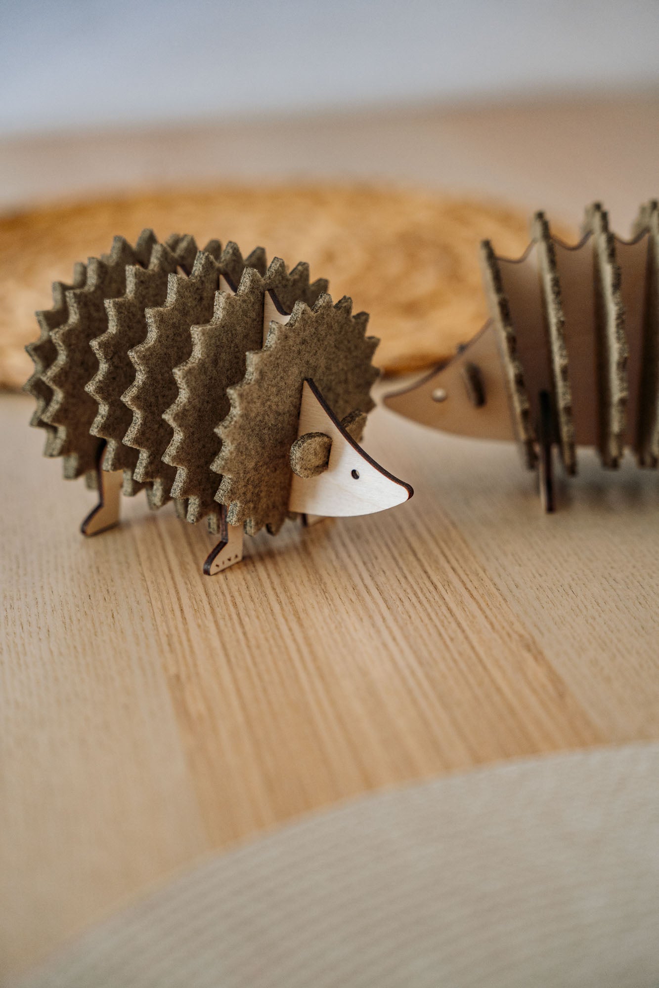 A small, decorative wooden hedgehog coaster holder made from layered, textured materials stands on a light wooden surface, with another similar figurine blurred in the background—perfect for adding charm to your home decor.