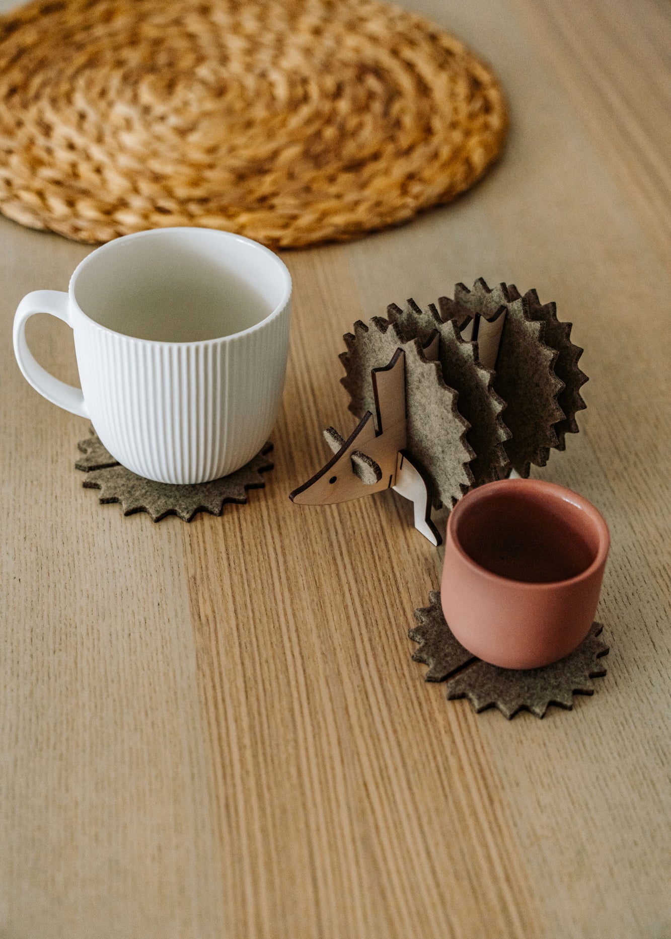 A white mug and a pink cup sit on spiky felt coasters shaped like a hedgehog, adding playful home decor flair to a light wooden table with a woven placemat in the background.