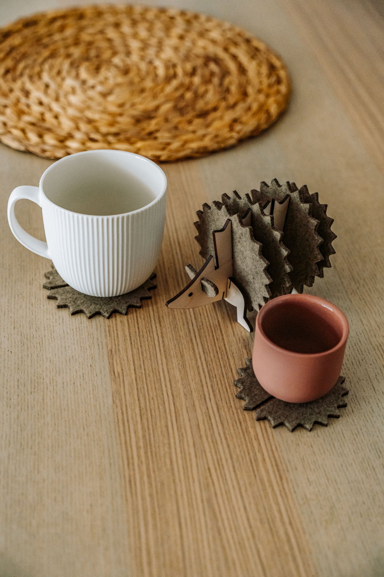 A white mug and a pink cup sit on spiky felt coasters shaped like a hedgehog, adding playful home decor flair to a light wooden table with a woven placemat in the background.