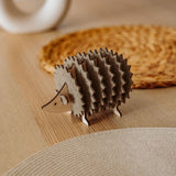 A small, decorative wooden hedgehog coaster holder sits on a wooden table with a woven placemat and a round, white ceramic object in the background, adding charm to your home decor.