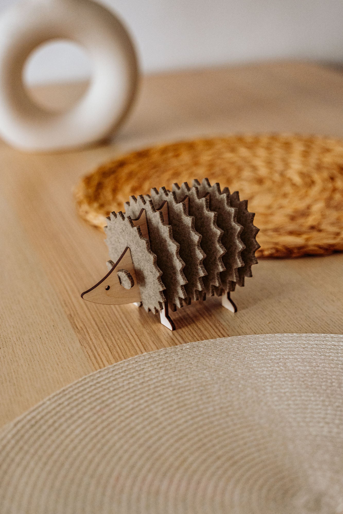 A small, decorative wooden hedgehog coaster holder sits on a wooden table with a woven placemat and a round, white ceramic object in the background, adding charm to your home decor.