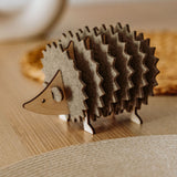 A charming wooden hedgehog coasters holder, perfect for home decor, sits on a light table with a woven placemat and blurred accents in the background.