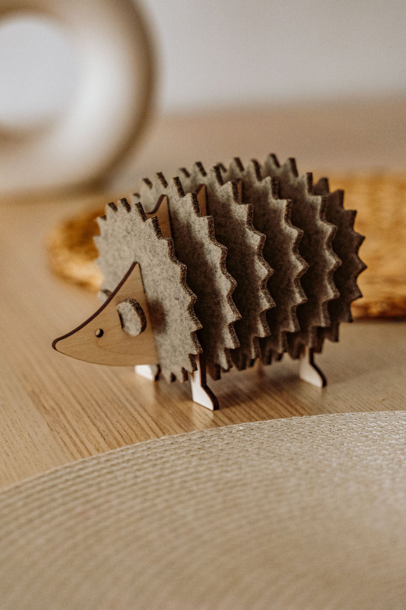 A charming wooden hedgehog coasters holder, perfect for home decor, sits on a light table with a woven placemat and blurred accents in the background.