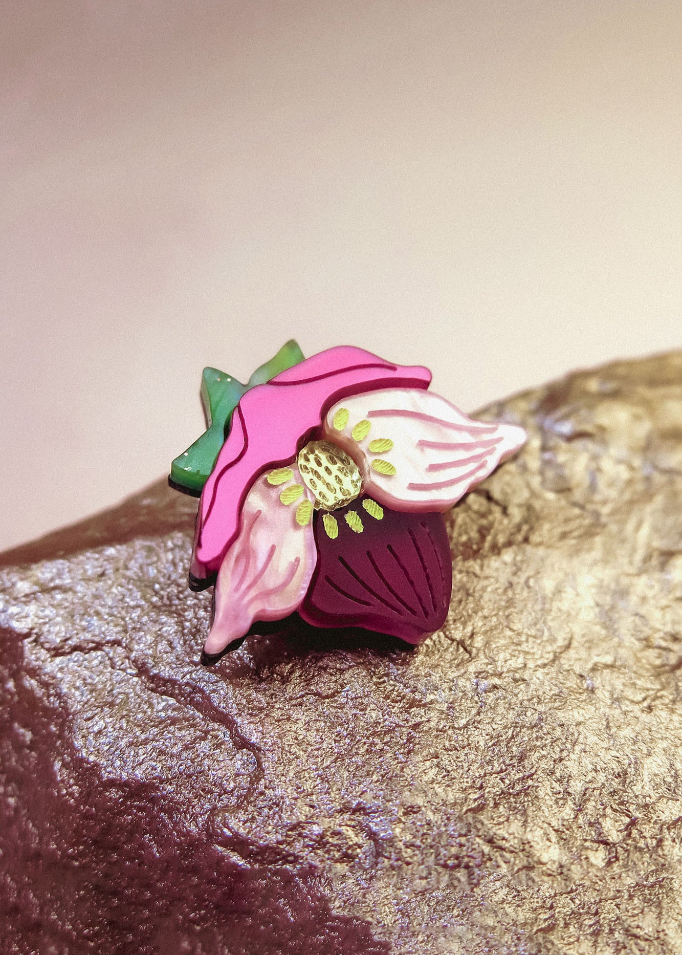 A colorful handmade lapel pin shaped like a purple hellebore flower, with pink petals, a green stem, and a yellow center, rests on a textured, metallic rock surface.