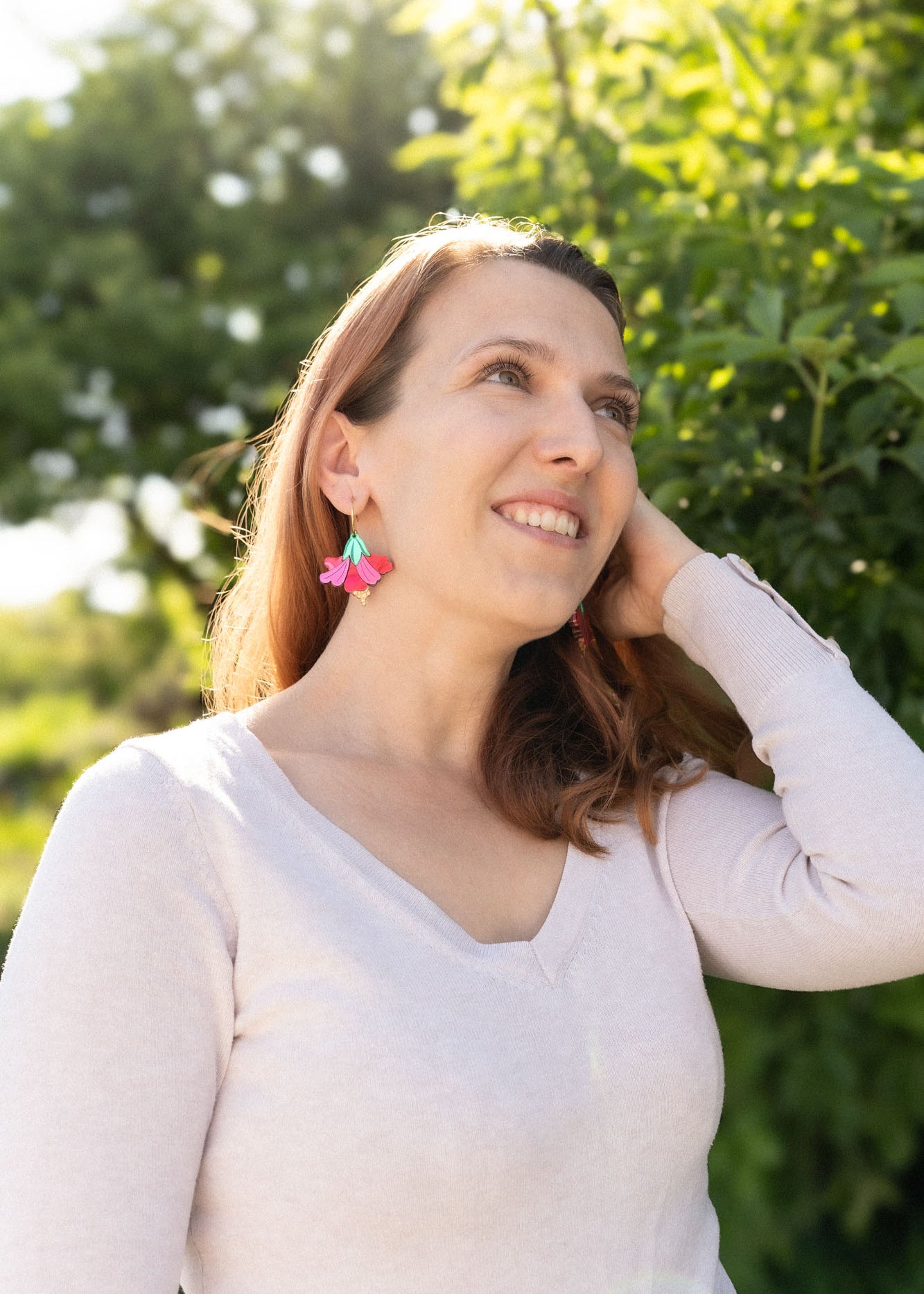 A woman with light brown hair smiles and looks upward, standing outdoors in sunlight. She wears a light long-sleeve top and handcrafted Hibiscus Earrings, with greenery and trees in the background.