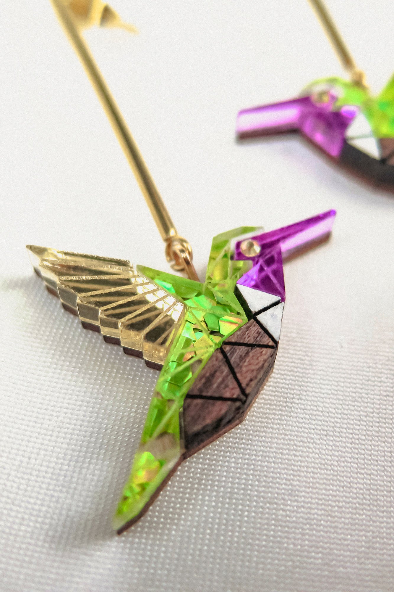 Close-up of handcrafted Hummingbird Earrings featuring gold, green, purple, and white geometric patterns, displayed on a white fabric background—a vibrant celebration of sustainable craftsmanship.