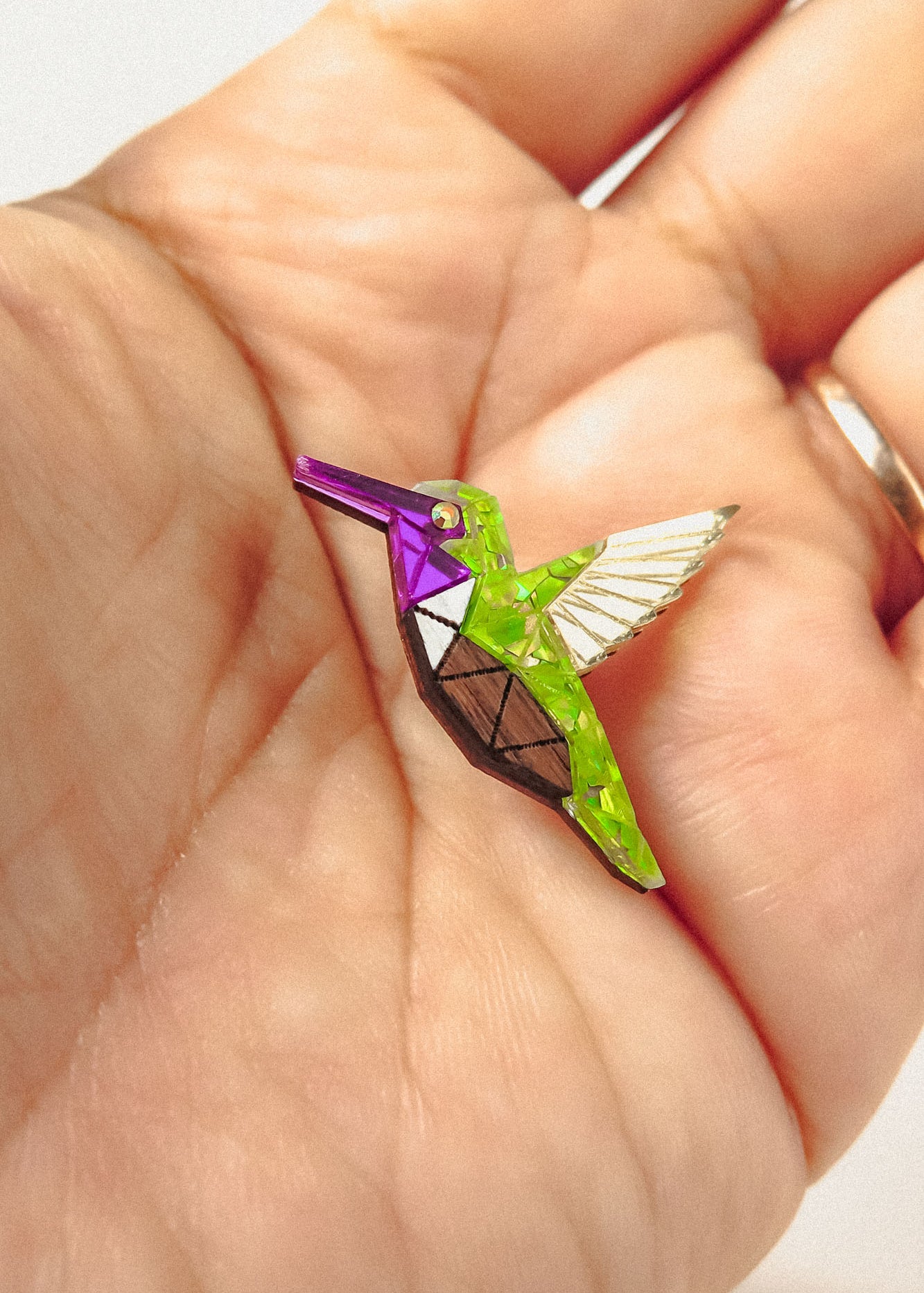 A hand holds a small, colorful Hummingbird Pin shaped like a geometric bird, featuring bright green, purple, and metallic segments with white and brown details—a nature-inspired design celebrating vibrant style.