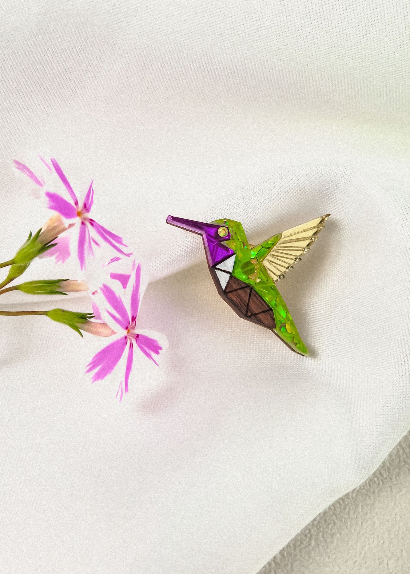 A colorful Hummingbird Pin with green, purple, brown, and gold sections rests on white fabric next to a sprig of pink flowers, showcasing nature-inspired design and timeless charm.
