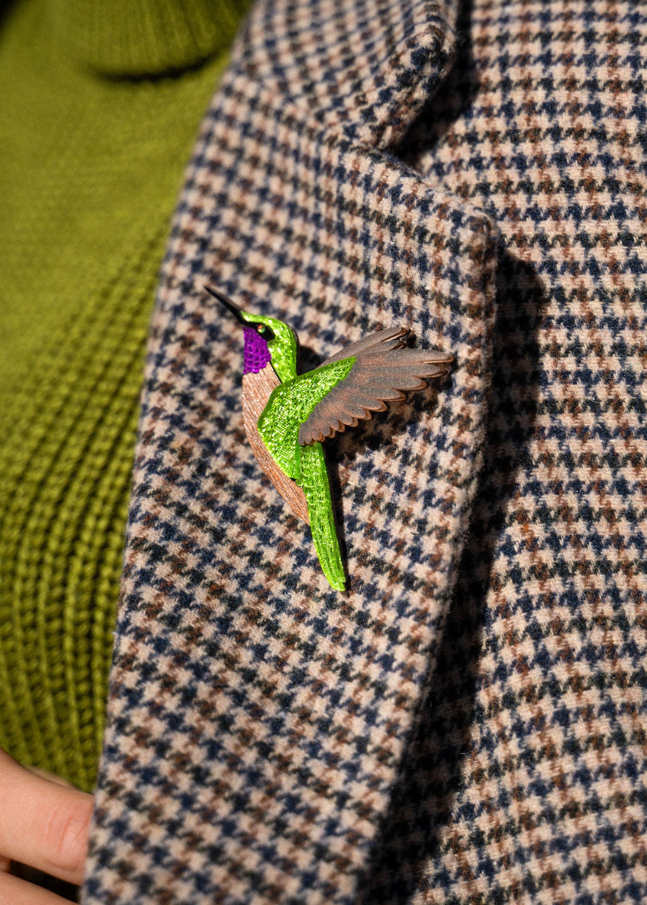 A close-up of a handcrafted hummingbird brooch, made from premium materials, pinned to a houndstooth-patterned coat with a green turtleneck sweater visible underneath.