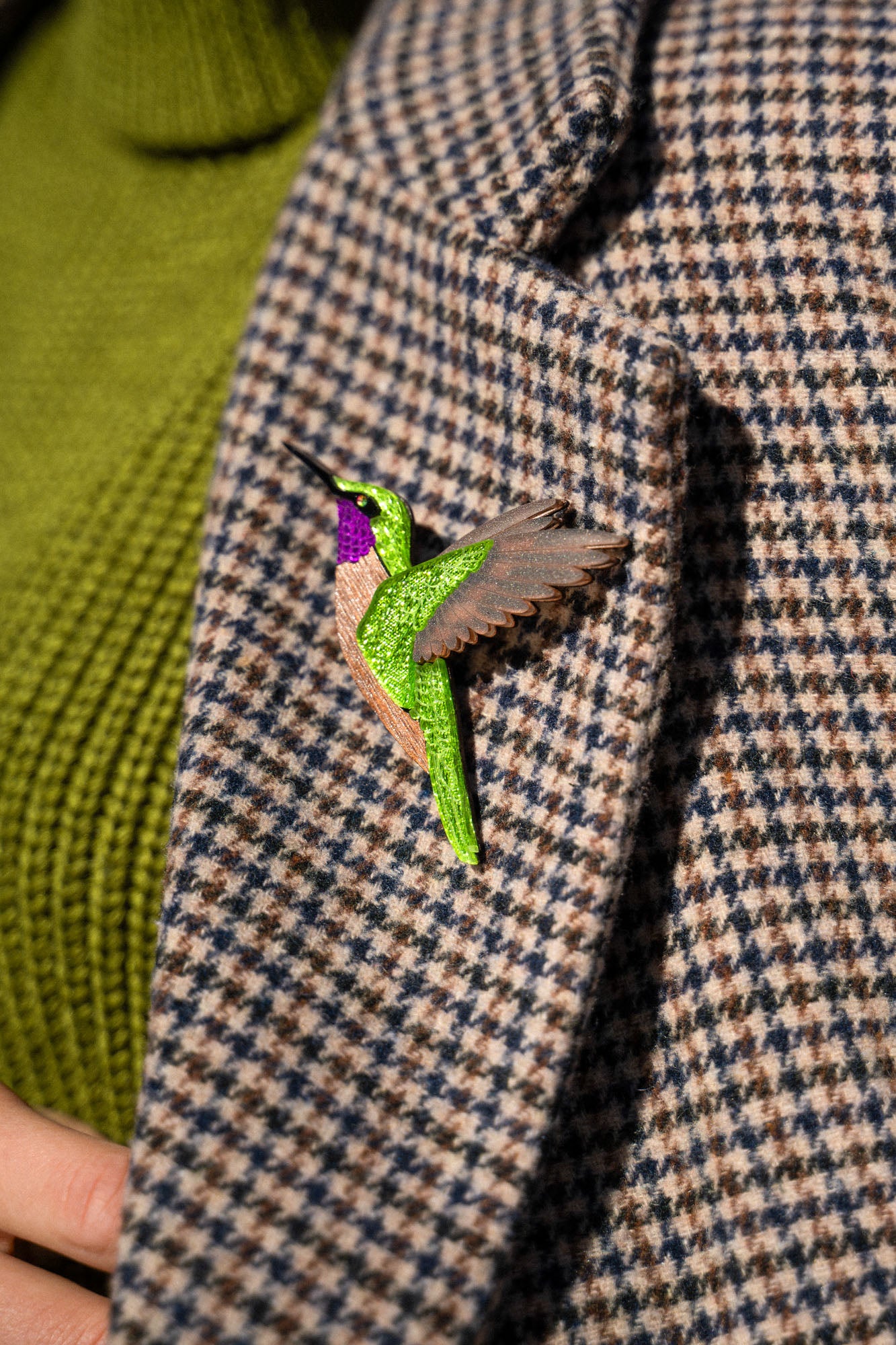 A close-up of a handcrafted hummingbird brooch, made from premium materials, pinned to a houndstooth-patterned coat with a green turtleneck sweater visible underneath.
