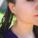 A close-up of a woman with closed eyes and braided hair, wearing handcrafted Hummingbird Earrings with pink details, set against an outdoor background.