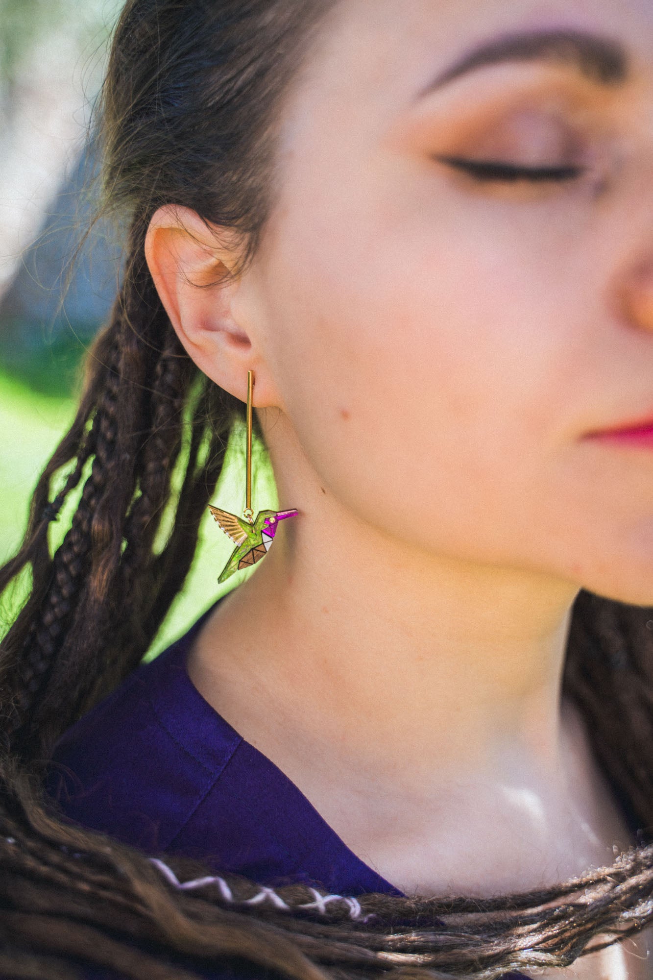 A close-up of a woman with closed eyes and braided hair, wearing handcrafted Hummingbird Earrings with pink details, set against an outdoor background.