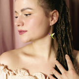 A woman with long brown dreadlocks and natural makeup wears an off-shoulder beige top, her hand to her chest. She showcases a colorful handcrafted hummingbird earring and stands before pink curtains, gazing thoughtfully to the side.