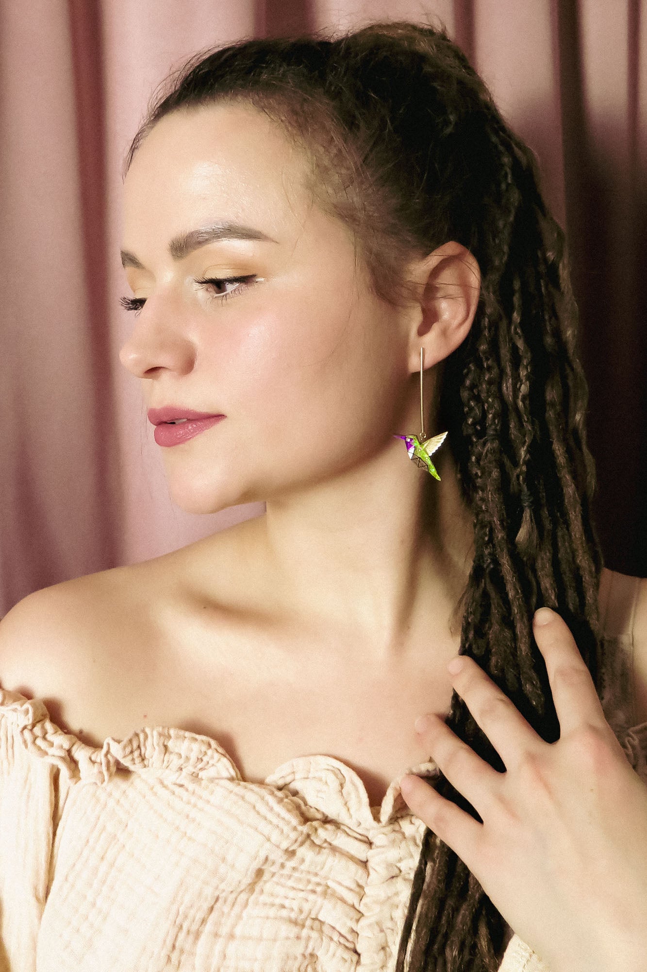 A woman with long brown dreadlocks and natural makeup wears an off-shoulder beige top, her hand to her chest. She showcases a colorful handcrafted hummingbird earring and stands before pink curtains, gazing thoughtfully to the side.