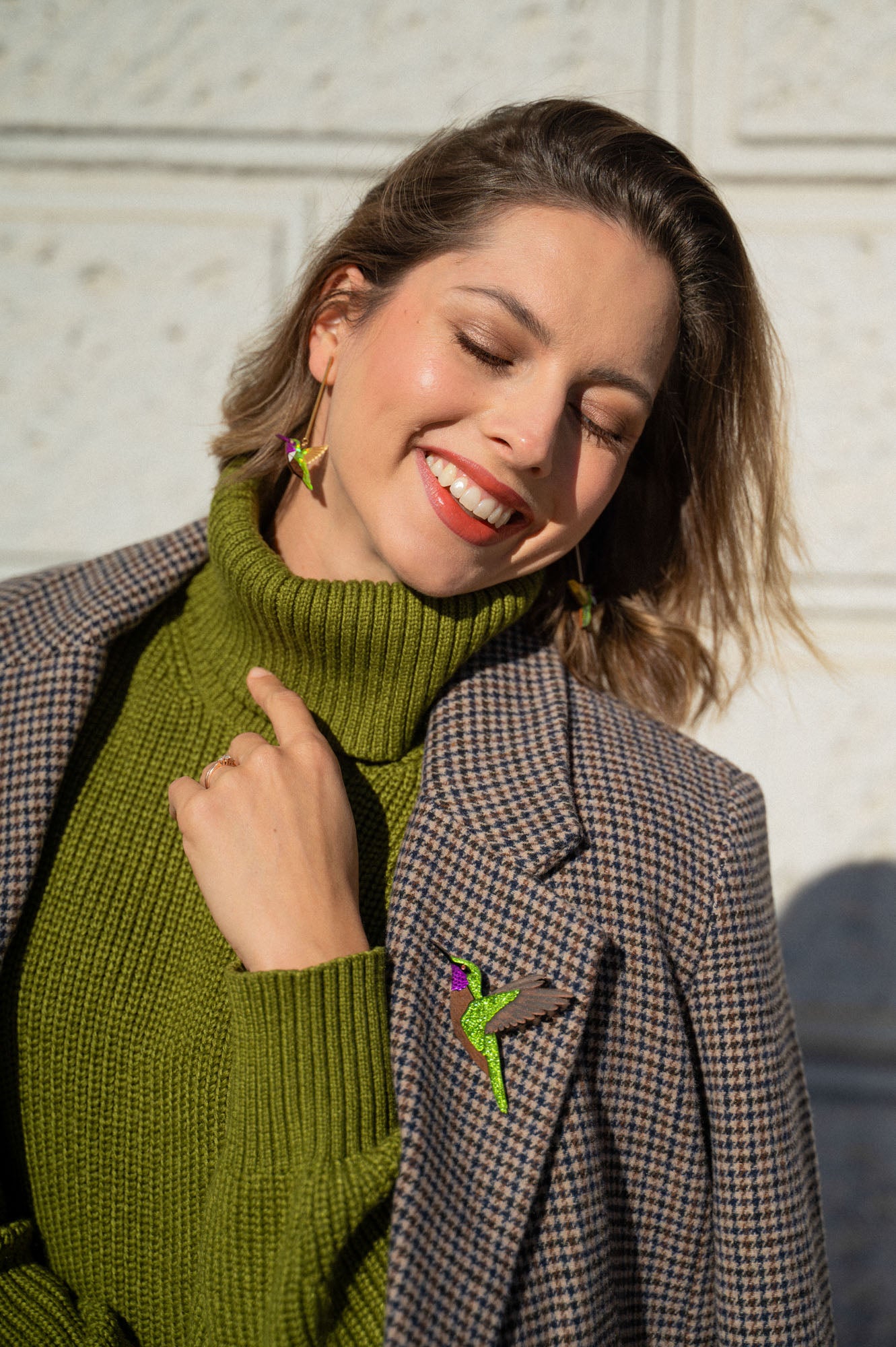 A smiling woman in a green turtleneck and plaid blazer stands in sunlight, eyes closed and head tilted. She wears bird-shaped earrings and a handcrafted Hummingbird Brooch, all made from premium materials.
