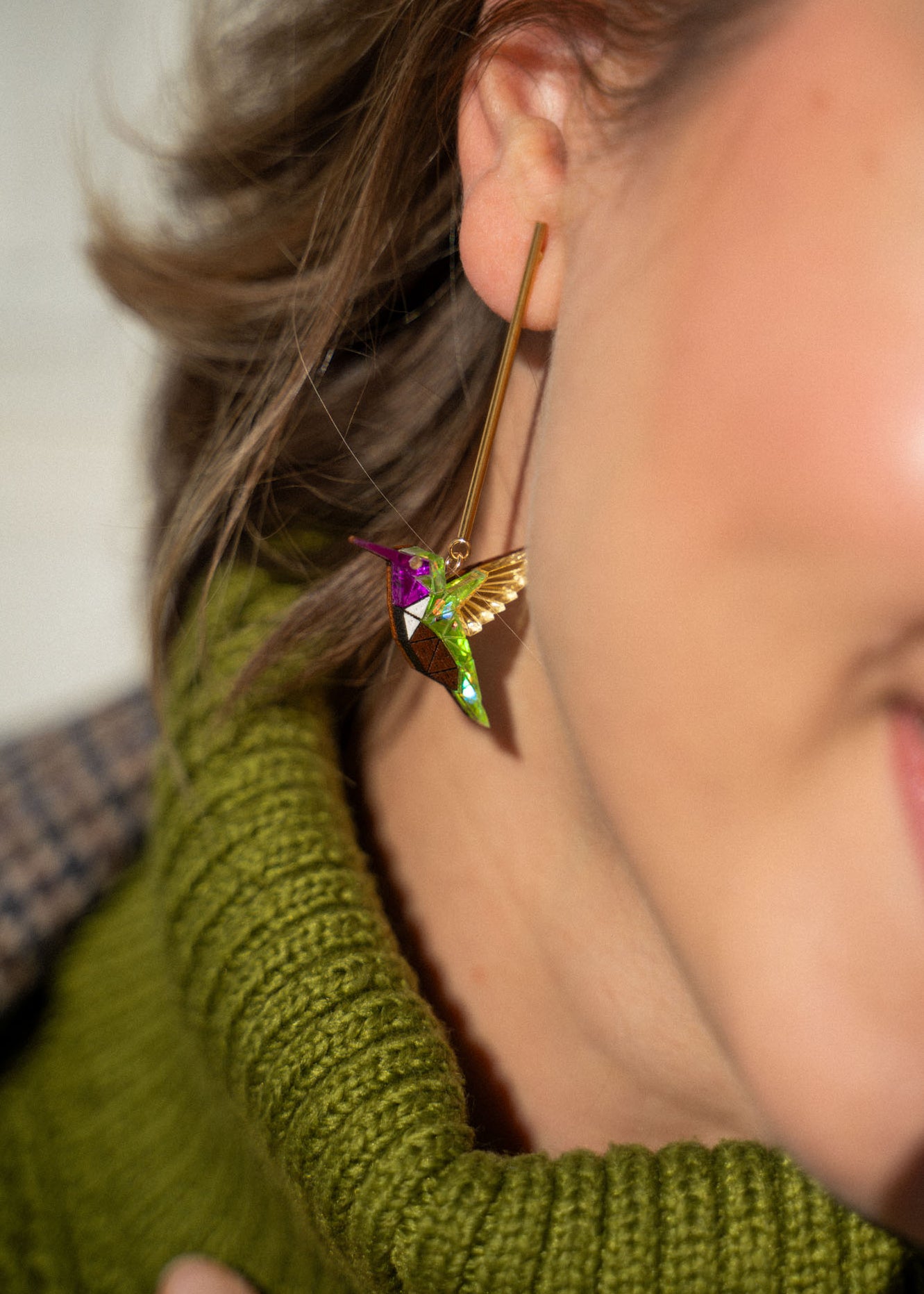 Close-up of a woman's ear wearing a long gold Hummingbird Earring, handcrafted with green, gold, and purple details. She pairs it with a green knitted turtleneck sweater, showcasing sustainable craftsmanship.