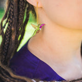 Close-up of a person wearing handcrafted Hummingbird Earrings, featuring green, yellow, and pink hues. The individual has braided hair and wears a purple top, highlighting the earring’s vibrant colors and sustainable craftsmanship.