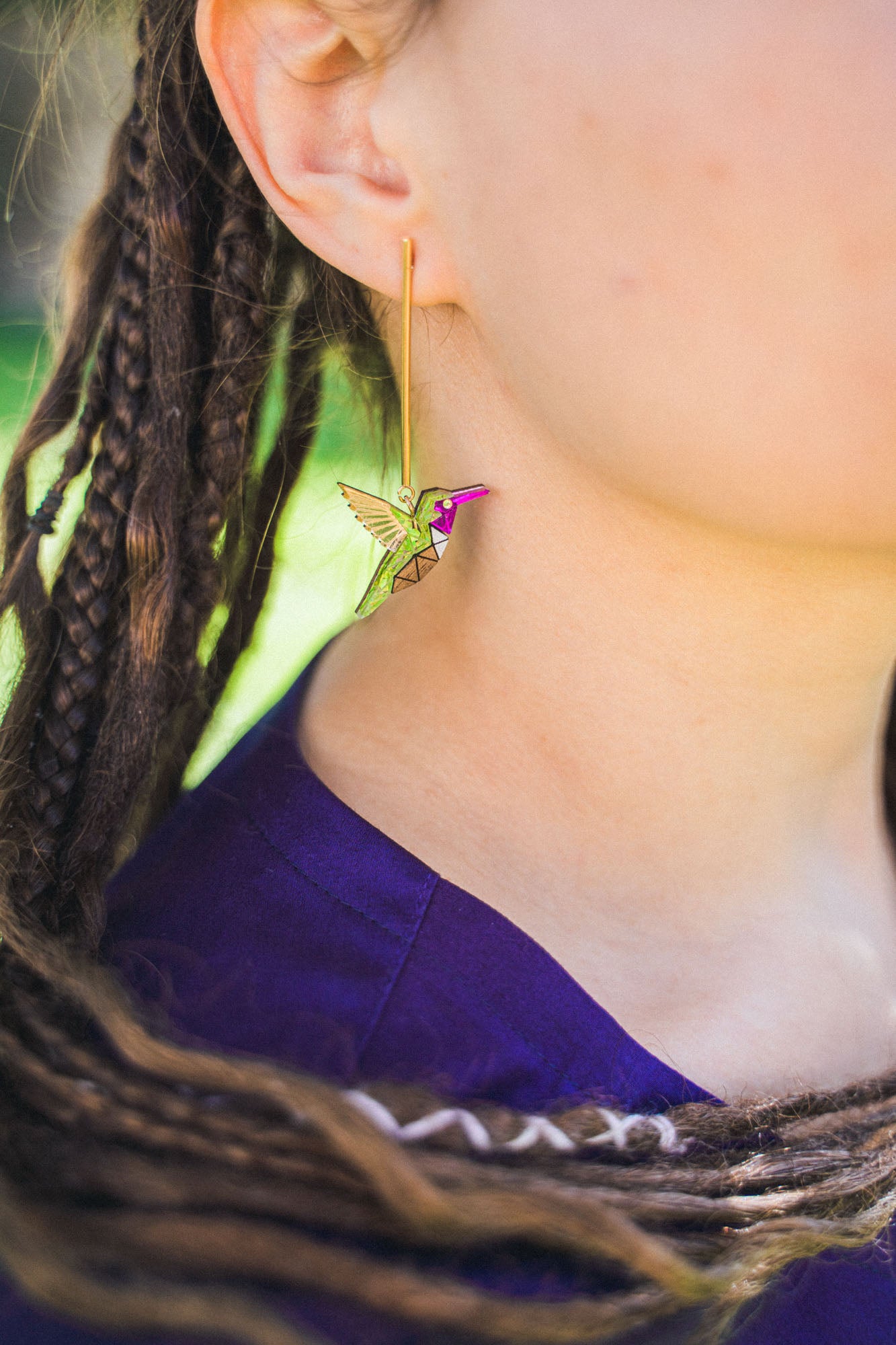 Close-up of a person wearing handcrafted Hummingbird Earrings, featuring green, yellow, and pink hues. The individual has braided hair and wears a purple top, highlighting the earring’s vibrant colors and sustainable craftsmanship.