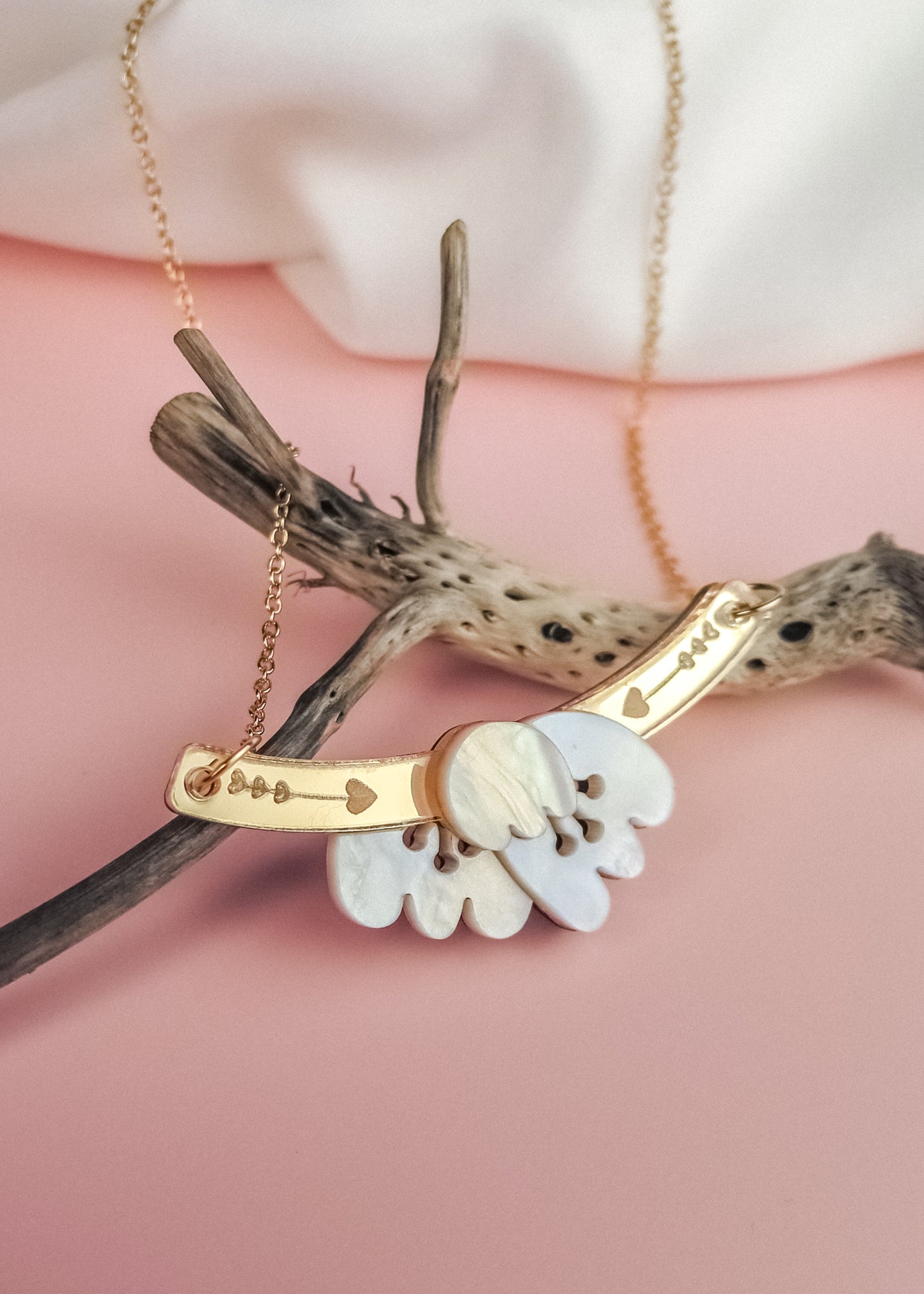 A handcrafted gold Tulip Necklace with engraved details rests on textured driftwood, featuring a white mother-of-pearl floral pendant, all set against a soft pink background for a touch of romantic charm.