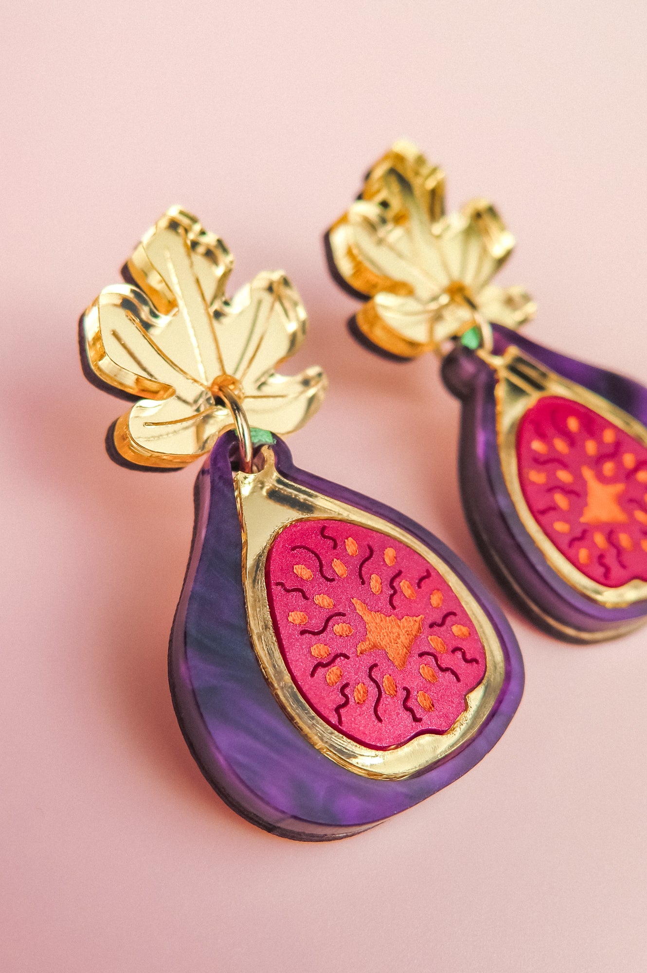 A close-up of handcrafted Fig Earrings shaped like figs, featuring gold mirror leaves at the top, vibrant purple edges, and a bright pink and orange fig center, set against a soft pink background.