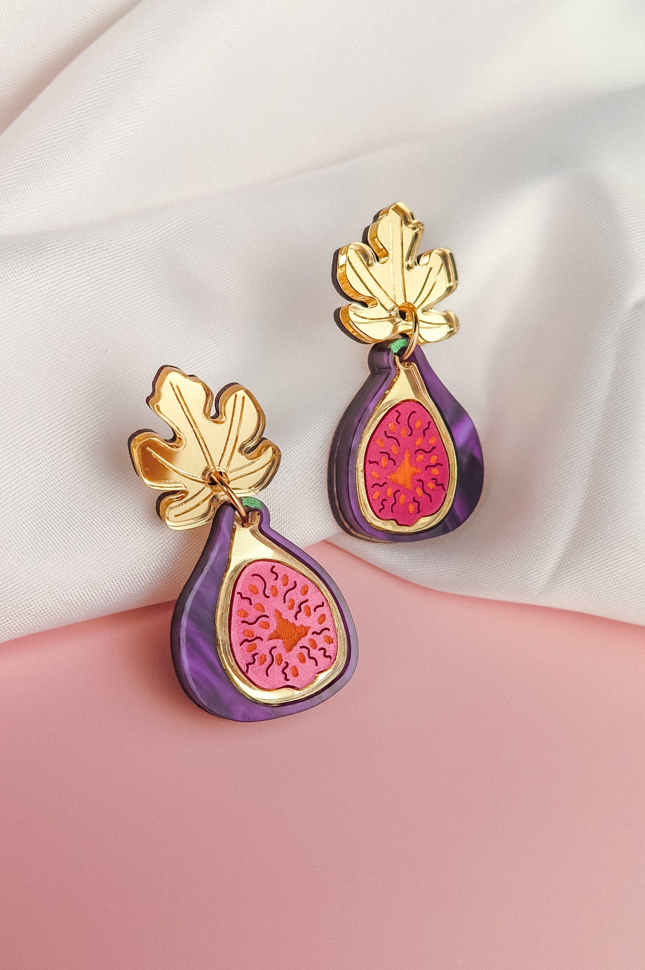 Handcrafted Fig Earrings shaped like figs with gold mirror leaves, featuring purple, pink, and gold details, displayed on a white and pink fabric background.