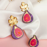 Three handcrafted Fig Earrings, featuring gold mirror leaves and vibrant purple and pink detailing, are displayed on white fabric and a pink surface, with dried foliage in the background.