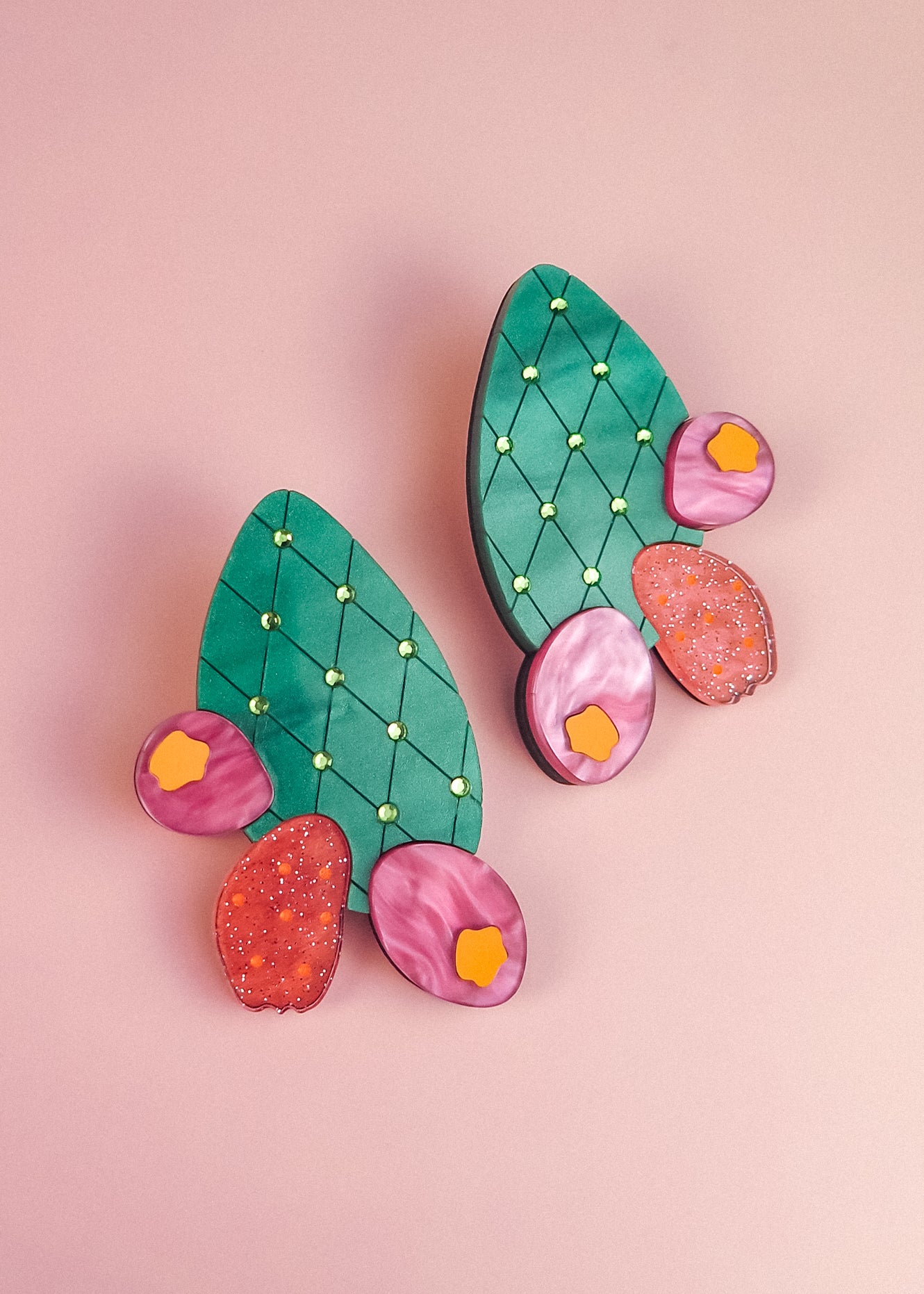 A pair of statement earrings shaped like green opuncia cactus pads with pink and orange flowers, decorated with small yellow dots, displayed on a light pink background.