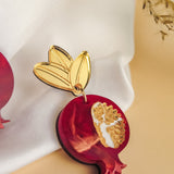 A handcrafted earring shaped like a red pomegranate with a golden leaf attached at the top, placed on white fabric with dried plant stems in the background, showcases bold designs and expert craftsmanship.