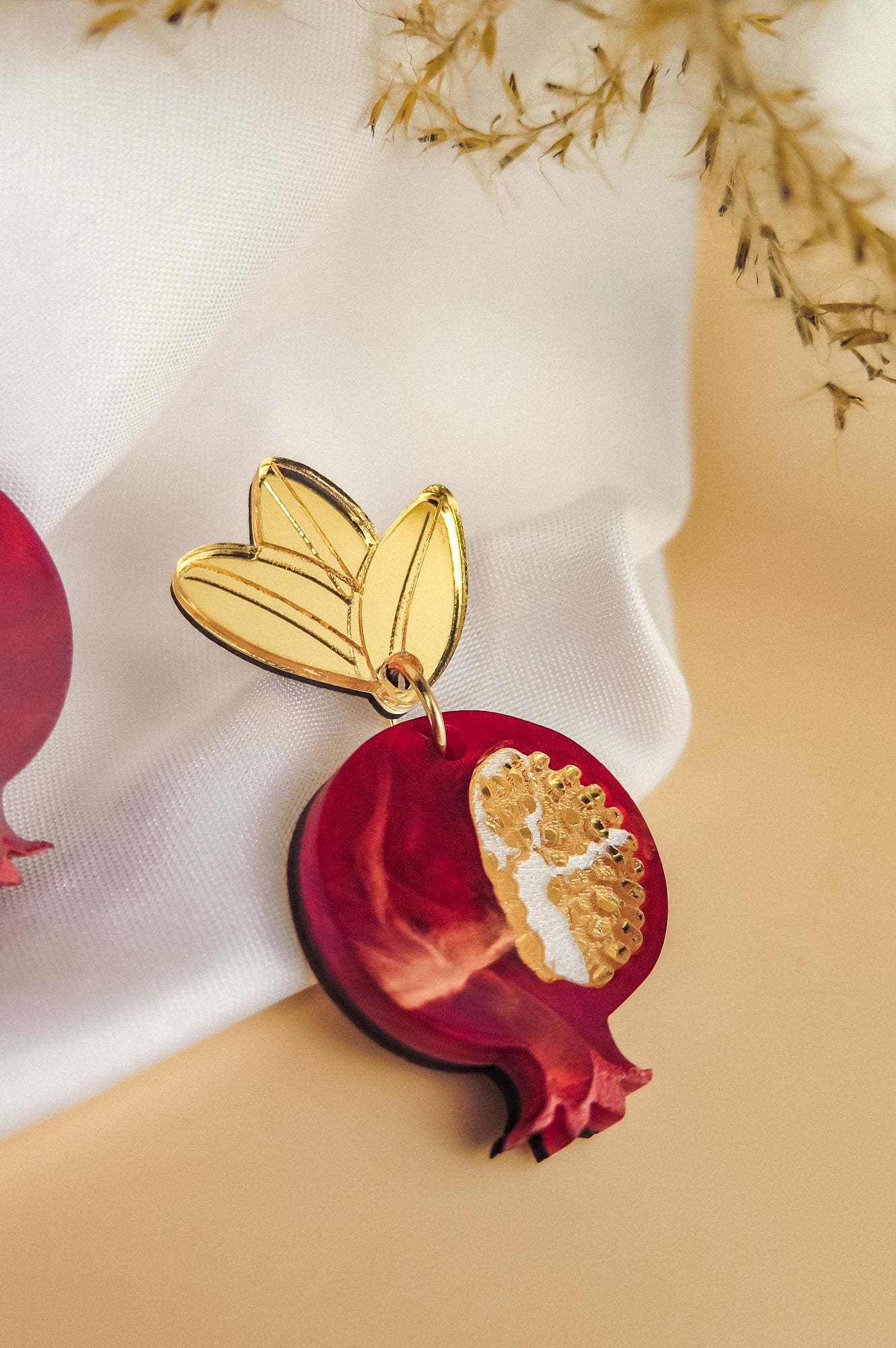 A handcrafted earring shaped like a red pomegranate with a golden leaf attached at the top, placed on white fabric with dried plant stems in the background, showcases bold designs and expert craftsmanship.
