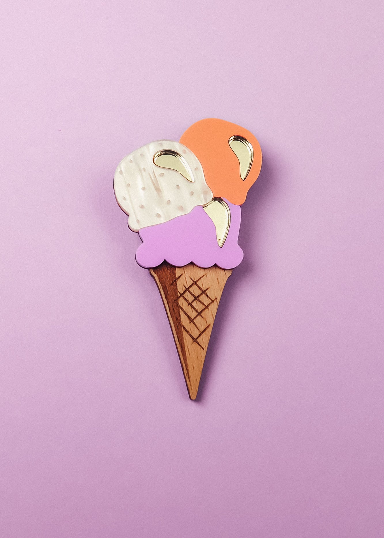 Handcrafted for a fun summer look, this Ice Cream Brooch features a wooden cone with three gold-accented scoops—white, purple, and orange—set against a pastel purple background.