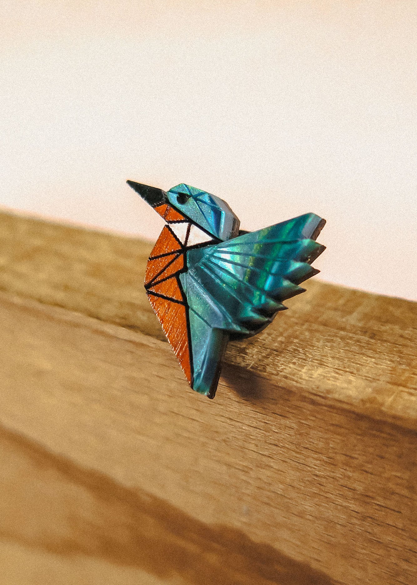 A colorful origami-style kingfisher pin brooch with blue, teal, and orange geometric sections, perched on the edge of recycled wood against a soft beige background—crafted from eco-friendly materials.