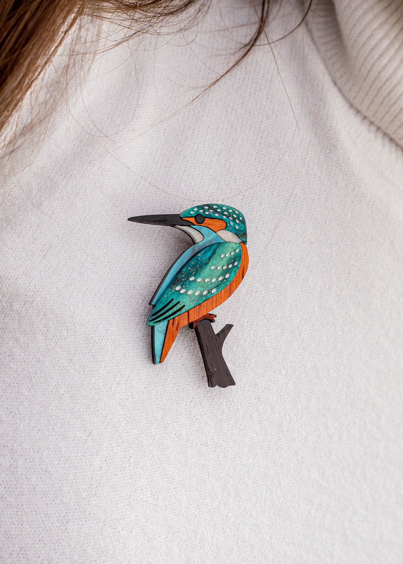 A handcrafted Kingfisher Brooch, featuring blue, orange, and white details, is pinned to a white textured sweater. Strands of brown hair are partially visible at the top of the image. Made using sustainable materials.