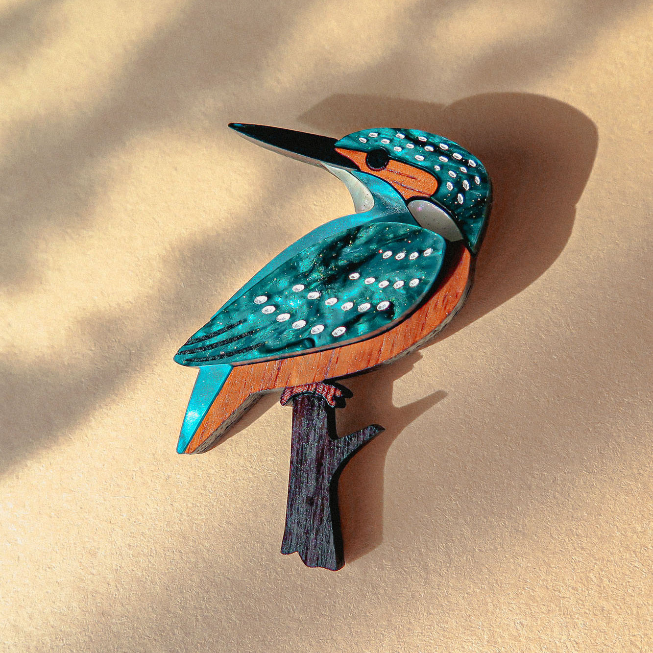 A colorful, handcrafted Kingfisher Brooch made from sustainable materials rests on a beige surface with soft shadows. The wooden brooch features striking blue, teal, white, and brown details.