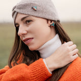 A woman wearing a grey headband with a kingfisher pin, a white turtleneck, and an orange sweater stands outdoors, looking to the side and touching her hair. The background is blurred and grassy—perfect for bird lovers seeking an eco-friendly accessory.