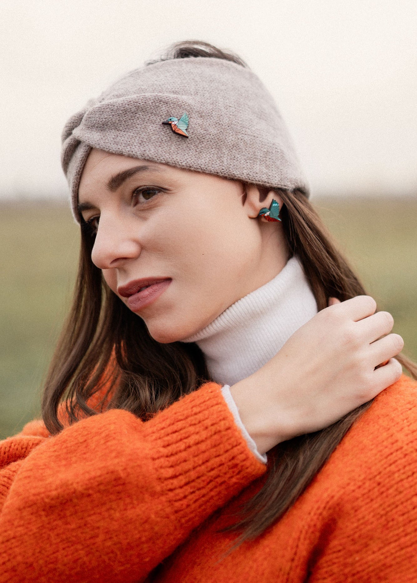 A woman wearing a grey headband with a kingfisher pin, a white turtleneck, and an orange sweater stands outdoors, looking to the side and touching her hair. The background is blurred and grassy—perfect for bird lovers seeking an eco-friendly accessory.